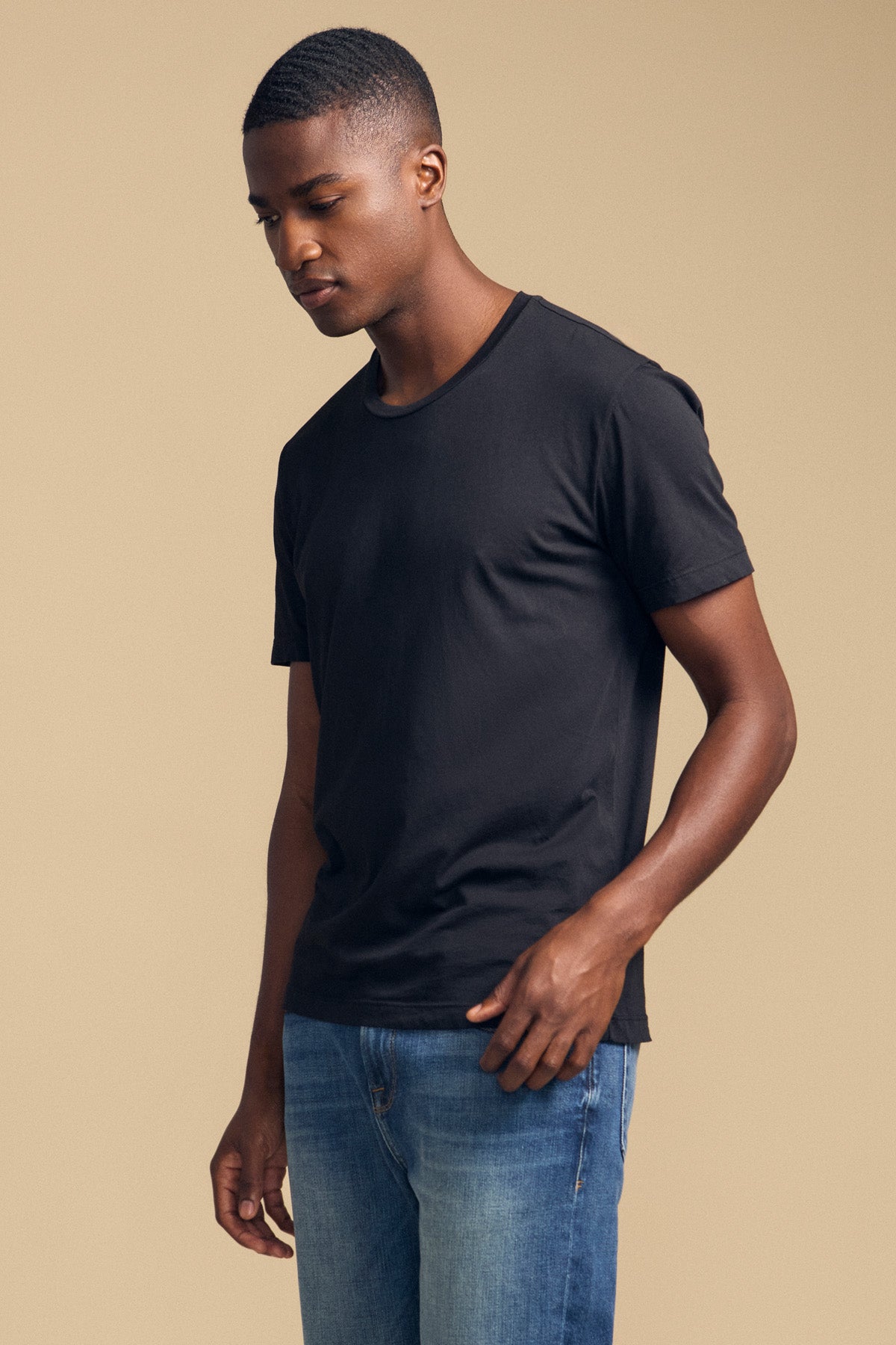 A man wearing the HOWARD TEE by Velvet by Graham & Spencer in plain black Pima jersey and blue jeans stands against a beige background, looking down with his left arm relaxed at his side.