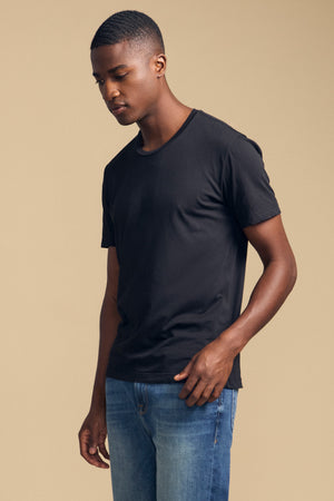 A man wearing the HOWARD TEE by Velvet by Graham & Spencer in plain black Pima jersey and blue jeans stands against a beige background, looking down with his left arm relaxed at his side.