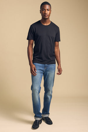 A man stands facing forward wearing the HOWARD TEE by Velvet by Graham & Spencer in black, paired with blue jeans and black shoes, against a neutral beige background.