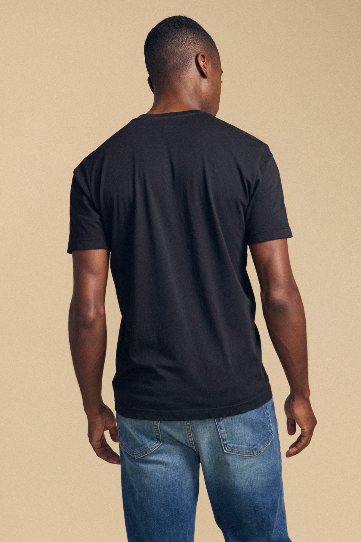A man stands facing away from the camera, wearing the Velvet by Graham & Spencer HOWARD TEE—a plain black short-sleeve t-shirt crafted from soft Pima jersey—paired with blue jeans against a beige background, showcasing its perfect fit for everyday comfort.