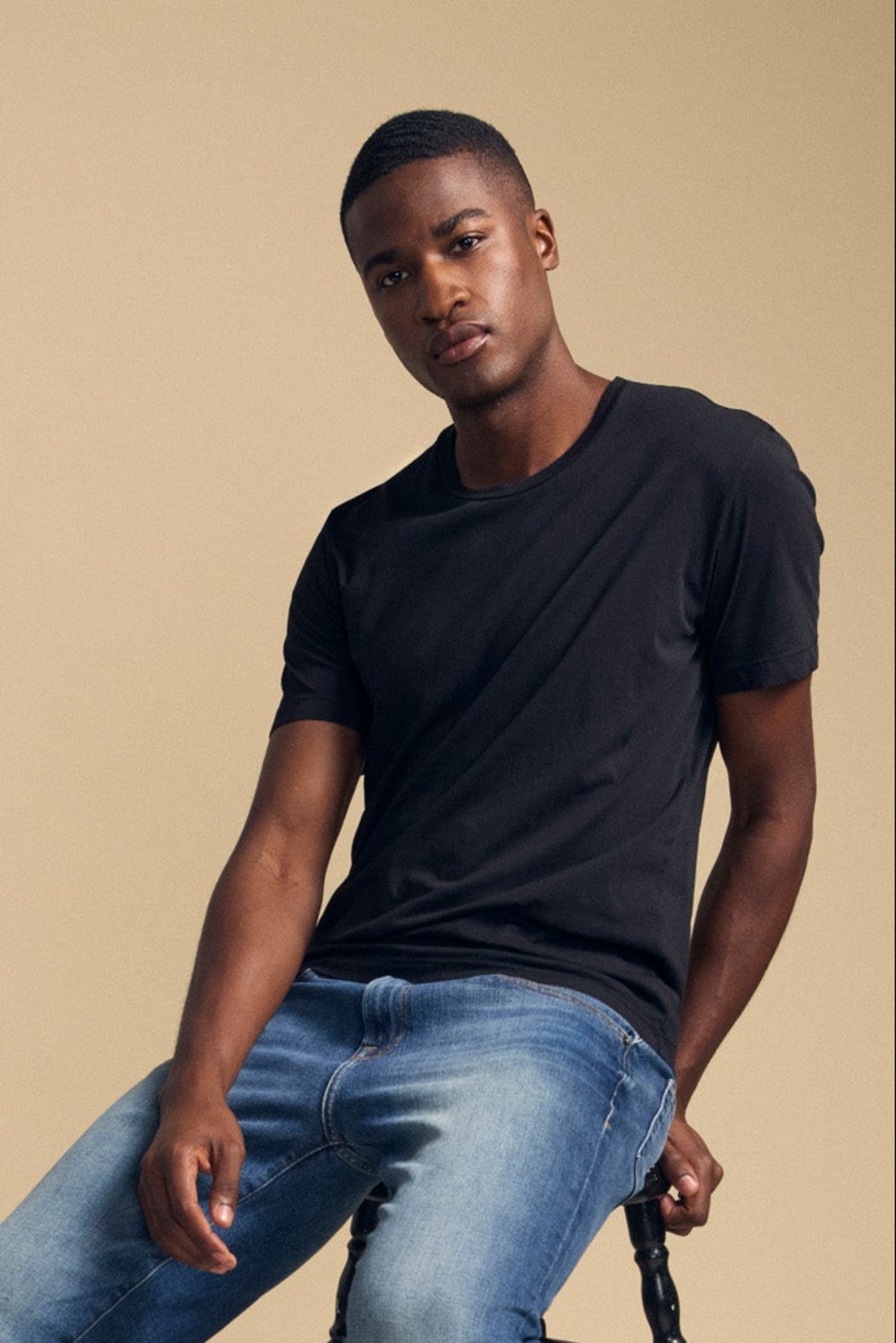 A man in the HOWARD TEE by Velvet by Graham & Spencer, paired with blue jeans and black loafers, sits on a wooden stool against a plain beige background, creating a vintage feel with his effortlessly perfect fit.-41774576107713