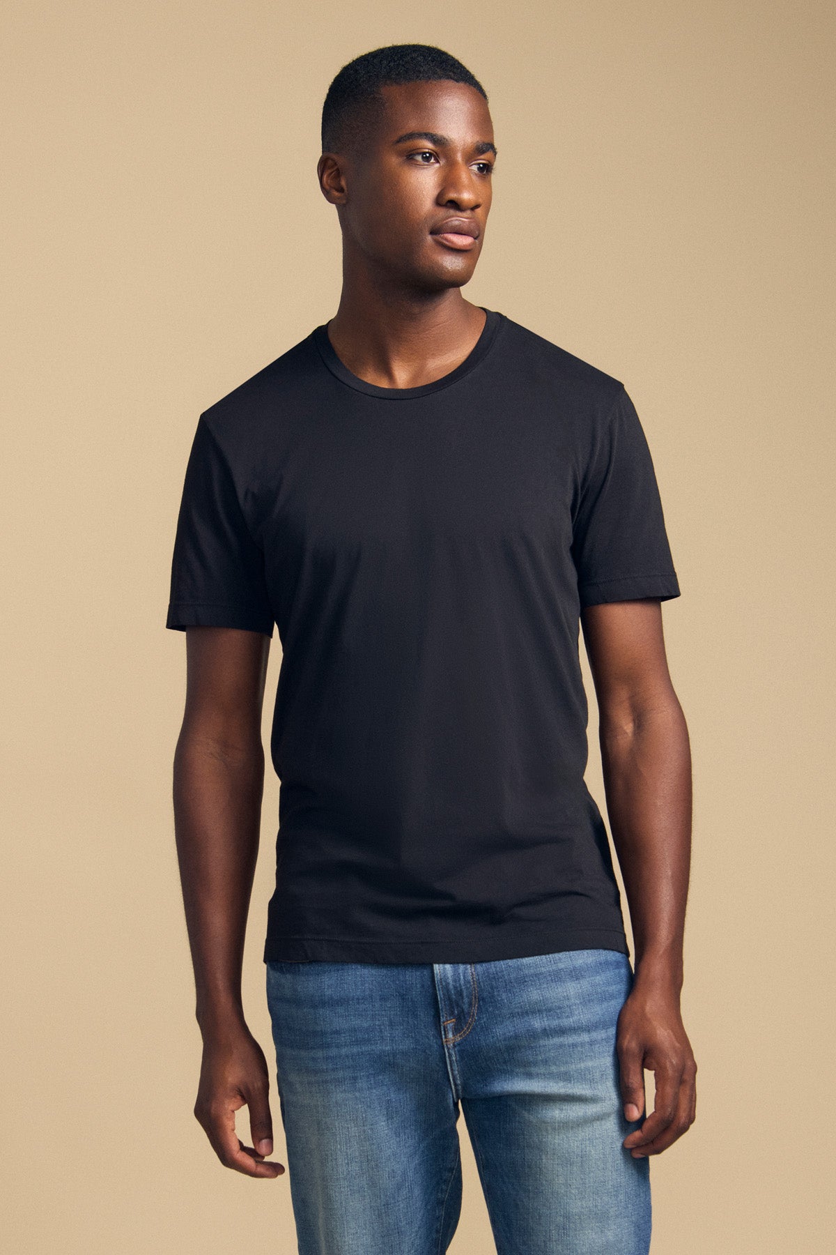 A person stands against a beige background wearing the HOWARD TEE by Velvet by Graham & Spencer—a plain black Pima jersey t-shirt paired with blue jeans—looking slightly to the side with a neutral expression. The tee’s perfect fit and vintage feel add subtle sophistication to the minimalist look.
