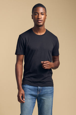 A man stands against a beige background, wearing the HOWARD TEE by Velvet by Graham & Spencer—a plain black Pima jersey t-shirt—paired with blue jeans. He looks at the camera with a neutral expression, and the outfit offers a vintage feel along with an impeccable fit.