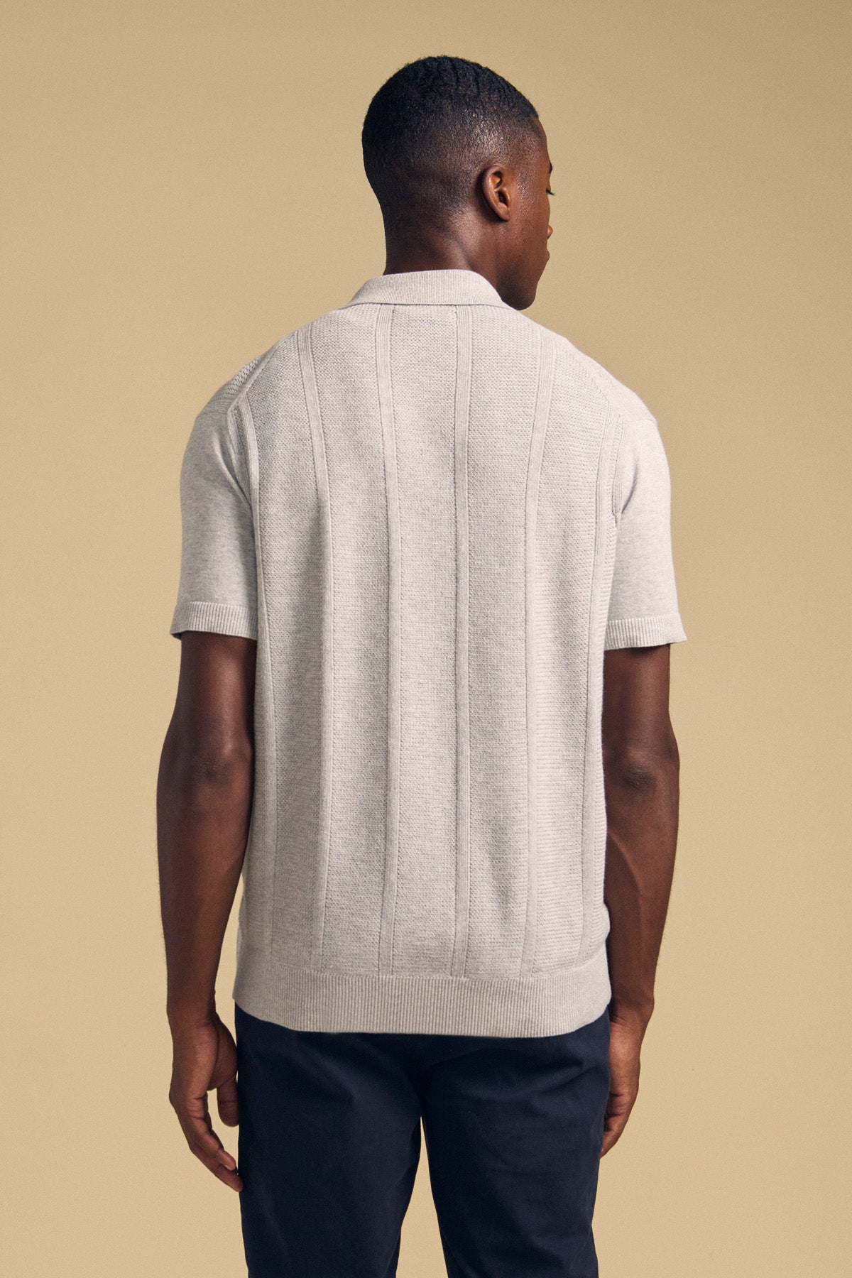 A man stands facing away, wearing the Velvet by Graham & Spencer LORENZO SWEATER POLO—a lightweight, breathable cotton-linen short-sleeve knit in light gray—paired with dark pants against a beige background.