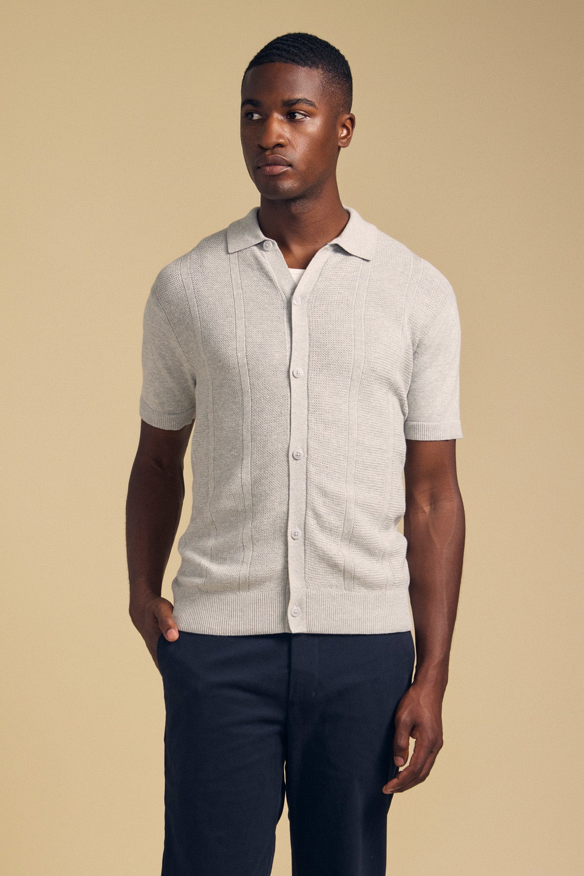 A man stands against a beige background wearing the LORENZO SWEATER POLO by Velvet by Graham & Spencer—a light gray, lightweight, breathable short-sleeve button-up sweater polo—paired with dark pants and has one hand in his pocket.