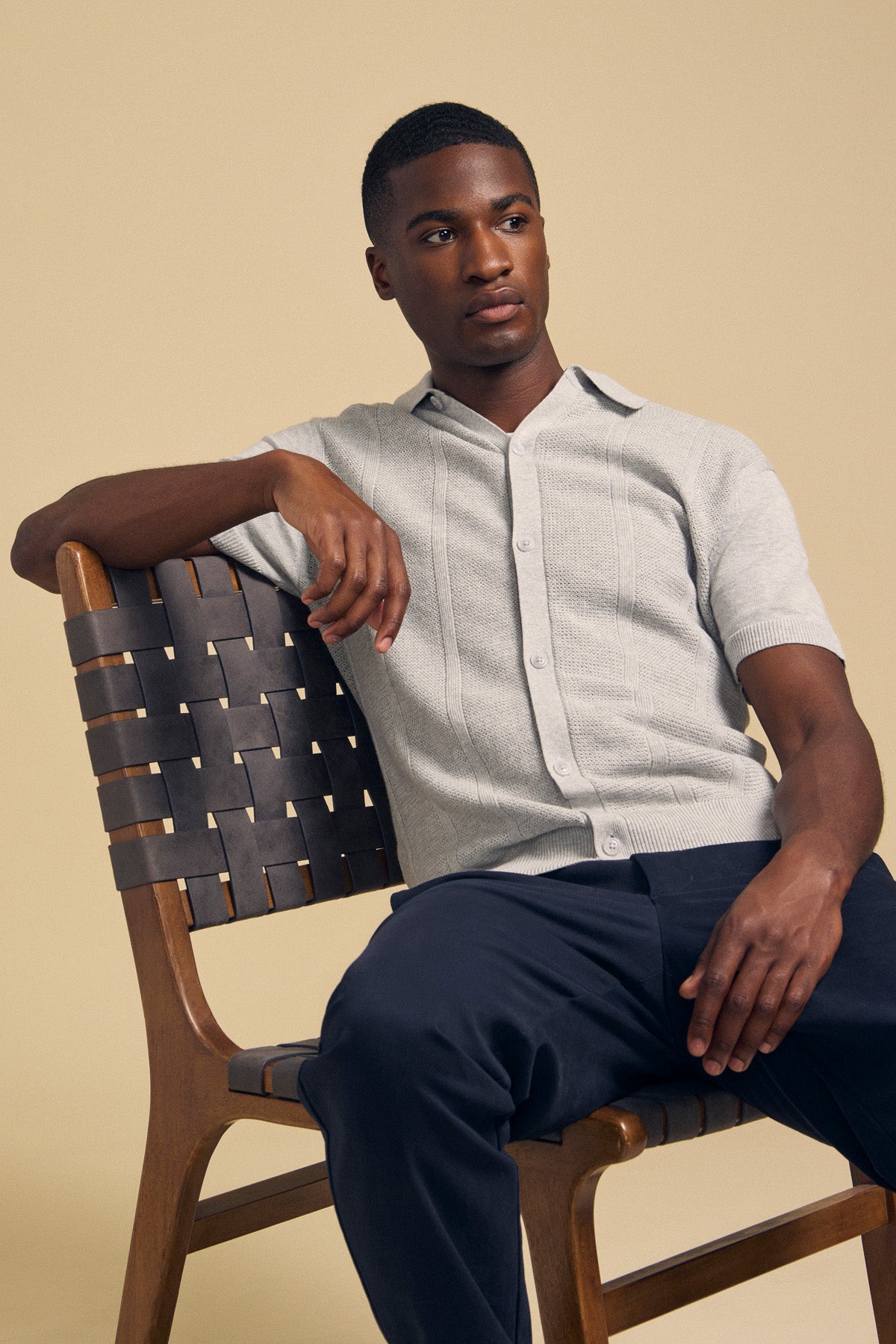A man sits on a wooden chair with a woven back, wearing the LORENZO SWEATER POLO from Velvet by Graham & Spencer—a lightweight, breathable textured cotton-linen polo—paired with dark pants. He looks off to the side against a plain beige background.-41859689414849