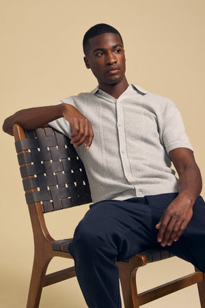 A man sits on a wooden chair with a woven back, wearing the LORENZO SWEATER POLO from Velvet by Graham & Spencer—a lightweight, breathable textured cotton-linen polo—paired with dark pants. He looks off to the side against a plain beige background.