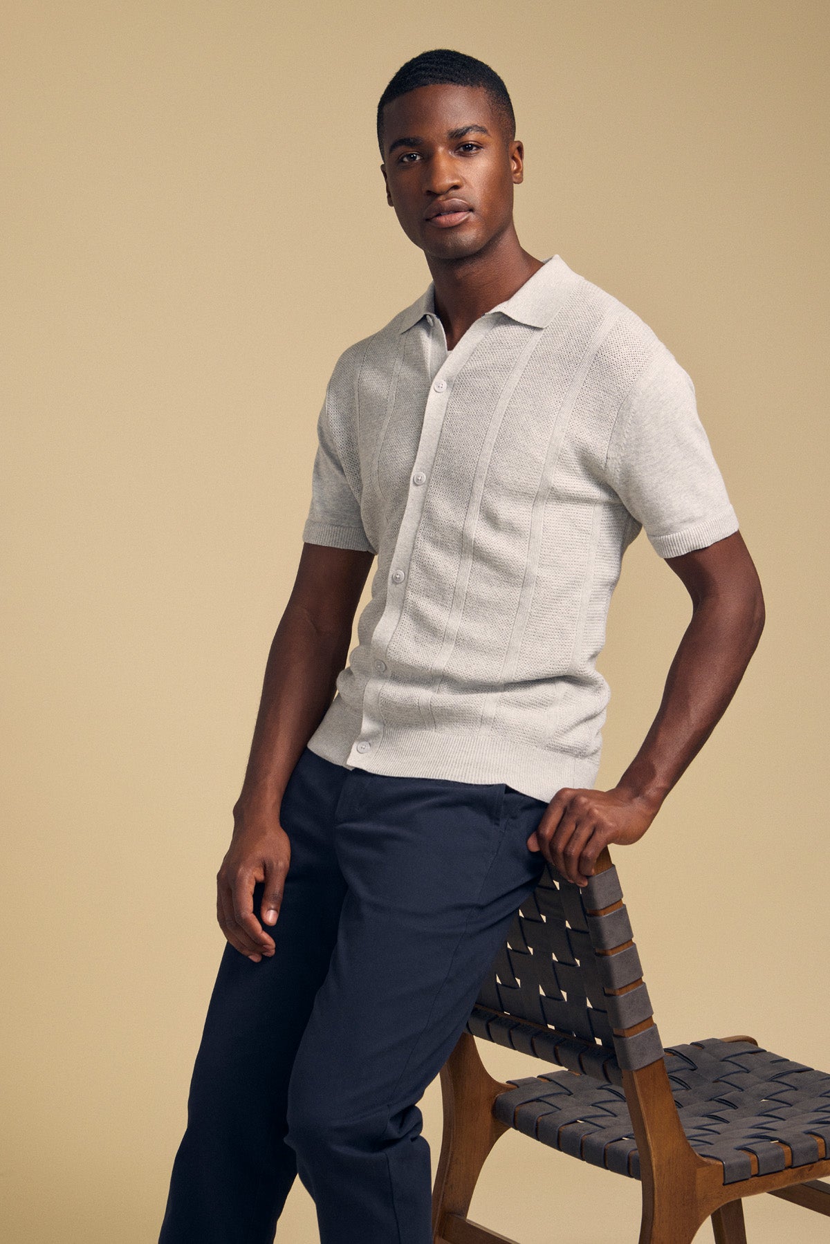 A man wearing the LORENZO SWEATER POLO by Velvet by Graham & Spencer in light gray, paired with dark pants, sits on the edge of a wooden chair against a beige background.