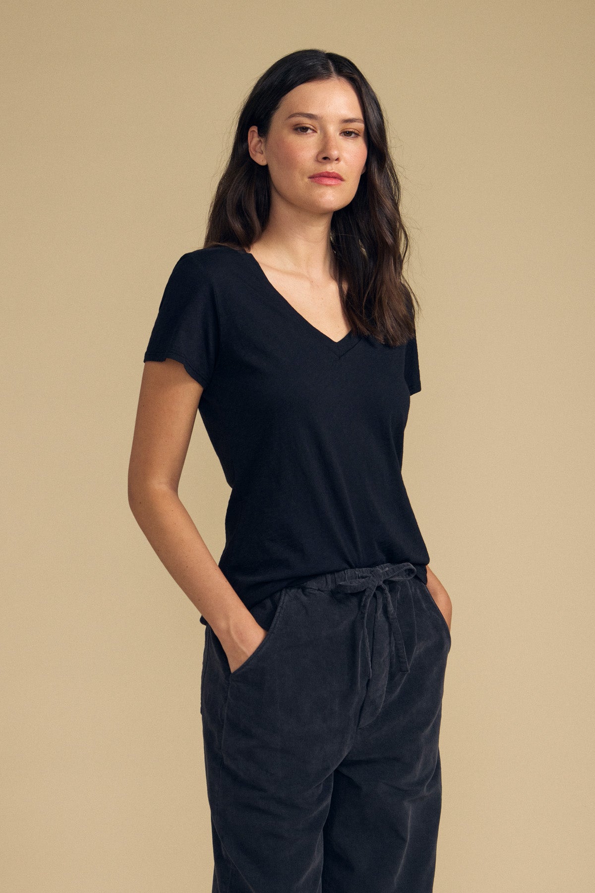 A woman with long dark hair stands against a beige background, wearing the LILITH TEE by Velvet by Graham & Spencer—a black cotton slub V-neck tee—paired with dark pants and her hands in her pockets for a flattering fit.