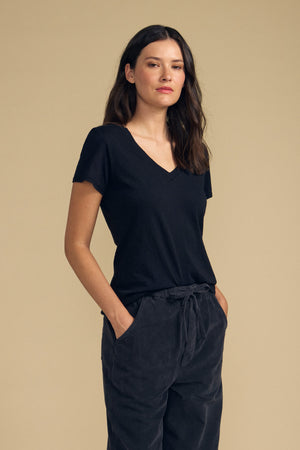 A woman with long dark hair stands against a beige background, wearing the LILITH TEE by Velvet by Graham & Spencer—a black cotton slub V-neck tee—paired with dark pants and her hands in her pockets for a flattering fit.