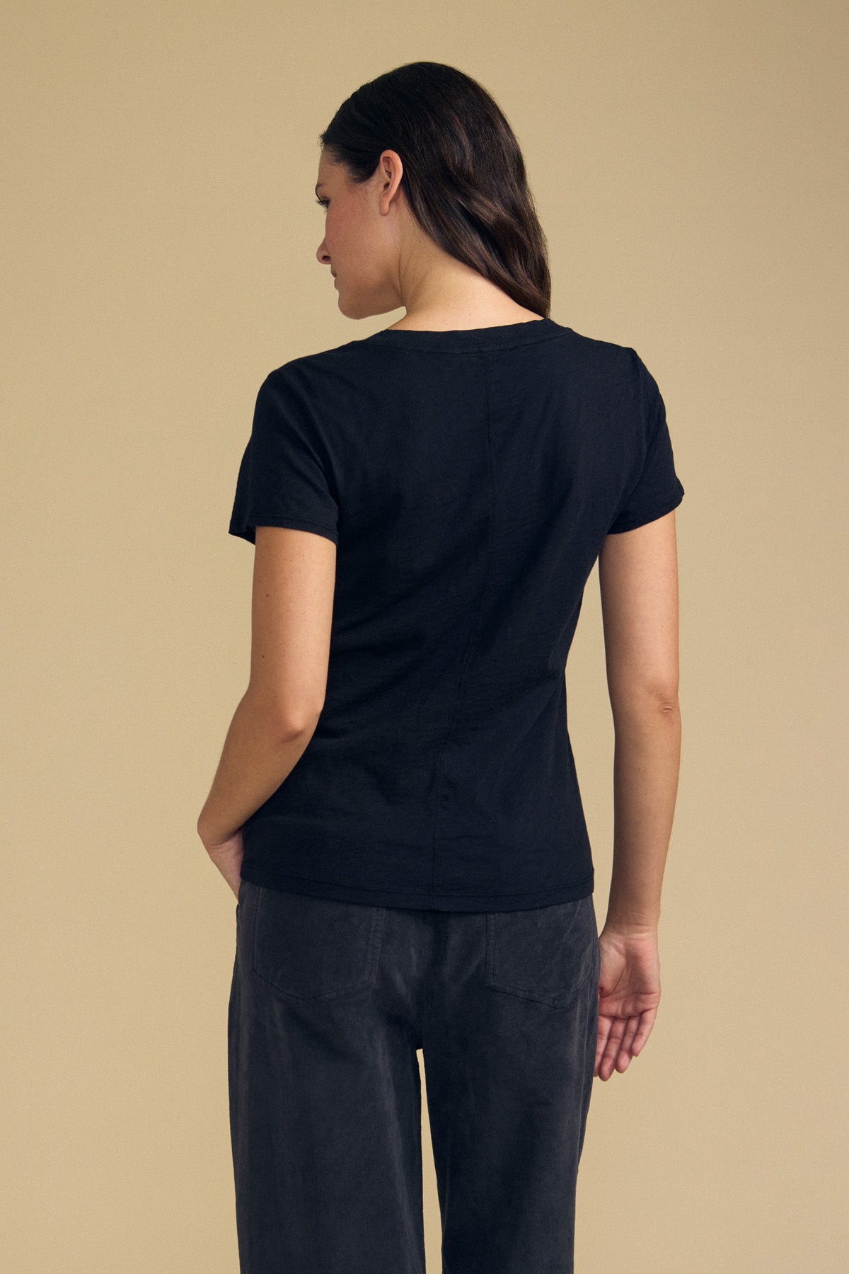 A person stands with their back to the camera, wearing Velvet by Graham & Spencer’s LILITH TEE—a short-sleeve black V-neck with a flattering fit—paired with dark pants, set against a plain beige background.-41925560959169