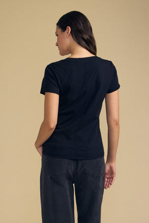 A person stands with their back to the camera, wearing Velvet by Graham & Spencer’s LILITH TEE—a short-sleeve black V-neck with a flattering fit—paired with dark pants, set against a plain beige background.