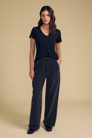 A woman stands against a beige background wearing the LILITH TEE by Velvet by Graham & Spencer—a black cotton slub V-neck tee with a flattering fit—paired with loose dark gray drawstring pants and black shoes, one hand in her pocket.
