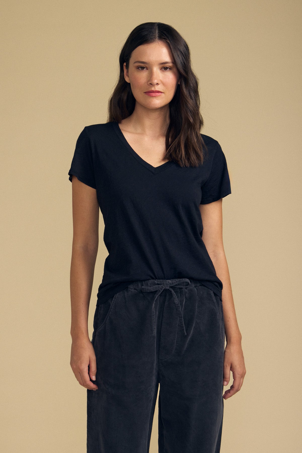 A woman wears the LILITH TEE by Velvet by Graham & Spencer—a black V-neck tee with a flattering fit—paired with dark drawstring pants, standing against a plain beige background.