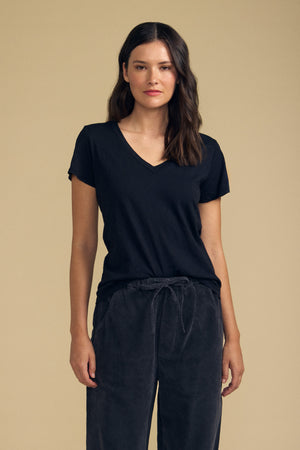 A woman wears the LILITH TEE by Velvet by Graham & Spencer—a black V-neck tee with a flattering fit—paired with dark drawstring pants, standing against a plain beige background.