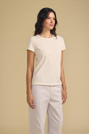 A woman stands against a beige background, wearing the ODELIA TEE by Velvet by Graham & Spencer—a cotton slub top featuring a classic crew neckline—paired with light-colored pants and looking forward with a neutral expression, effortlessly showcasing this wardrobe essential.
