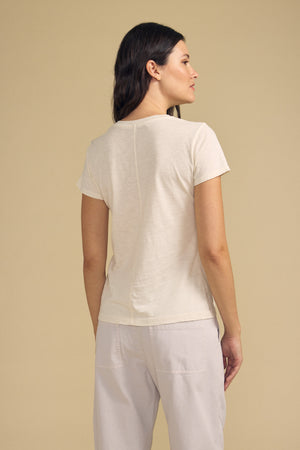A woman stands with her back turned, wearing the ODELIA TEE by Velvet by Graham & Spencer—a cotton slub tee with a classic crew neckline—paired with light pants against a plain beige background, embodying a true wardrobe essential.
