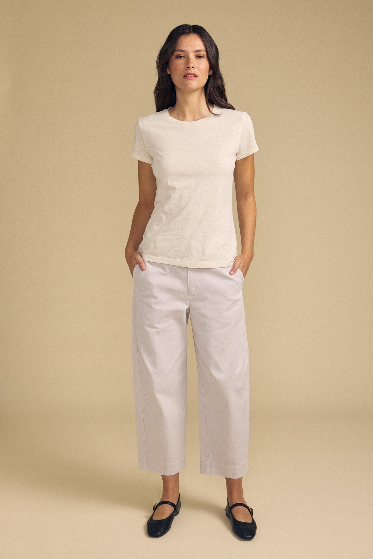 A woman stands against a beige background wearing the ODELIA TEE by Velvet by Graham & Spencer—a short-sleeve cotton slub tee with a classic crew neckline—paired with wide-leg light pants and black strap shoes, her hands resting in her pockets.