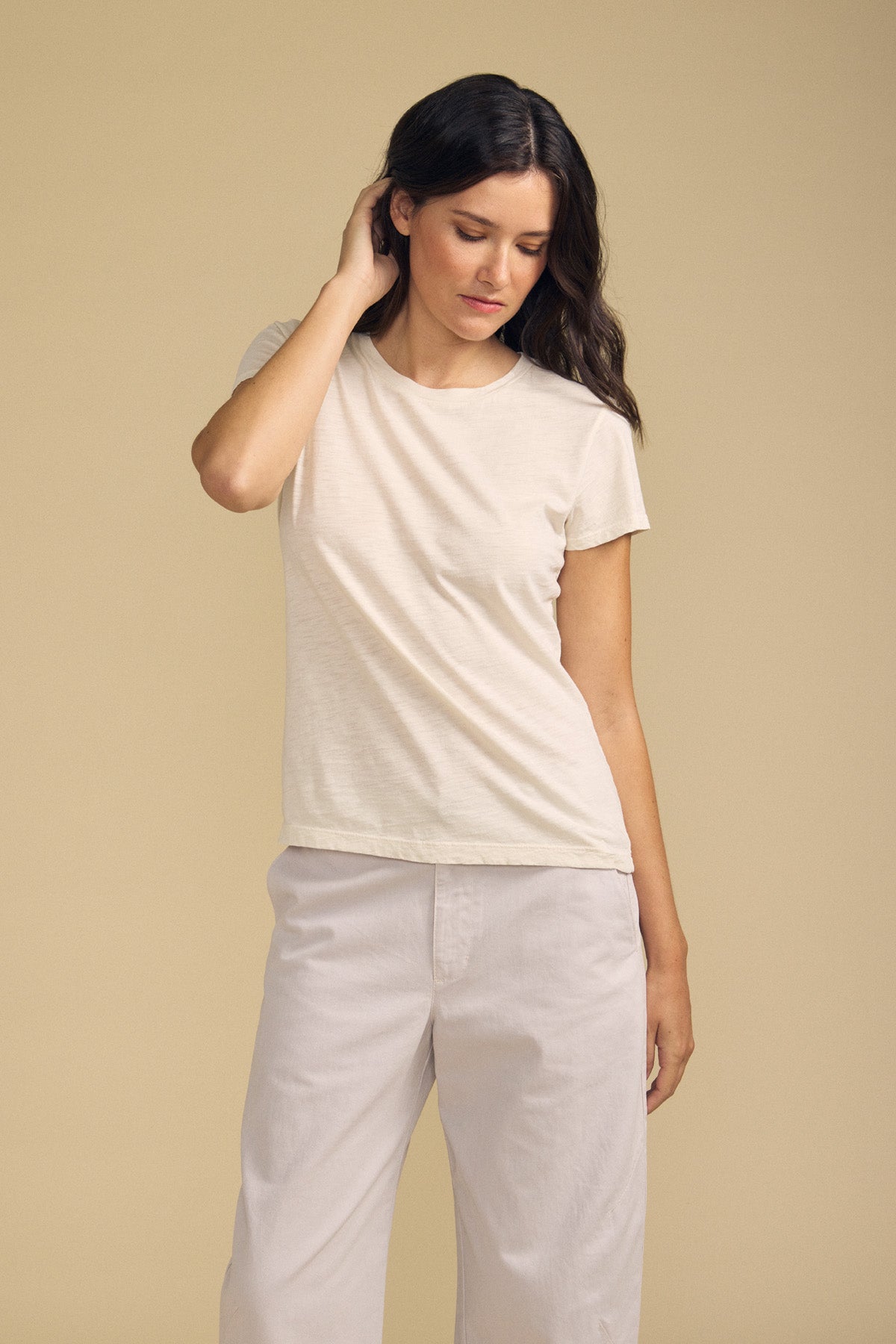 Against a beige background, a woman stands wearing the ODELIA TEE by Velvet by Graham & Spencer—a short-sleeve cotton slub essential featuring a classic crew neckline—paired with light-colored pants. She touches her neck with one hand and looks down.