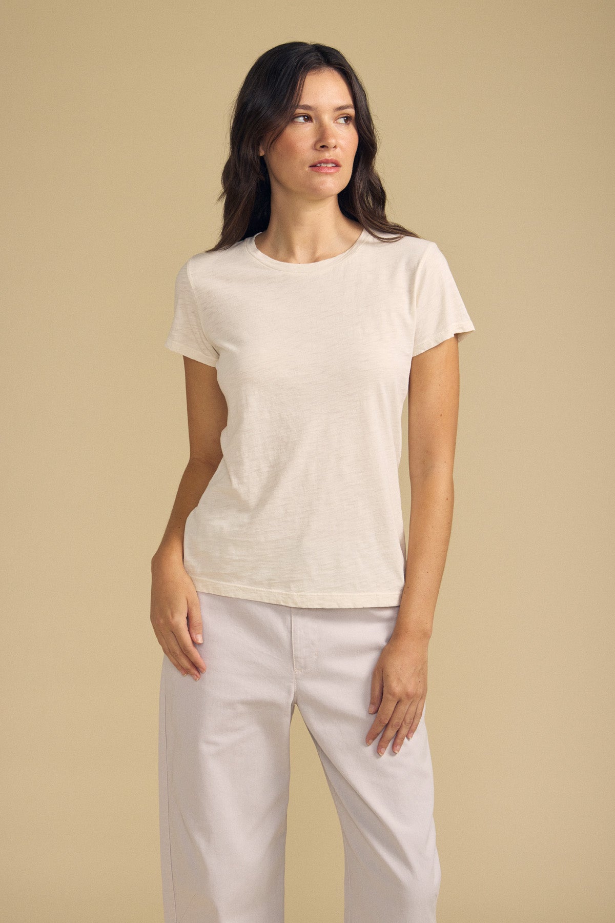 A woman wearing the ODELIA TEE by Velvet by Graham & Spencer—a short-sleeved beige cotton slub top with a classic crew neckline—pairs it with light pants as she stands against a plain beige background, gazing slightly to the side. This wardrobe essential adds understated elegance to any look.