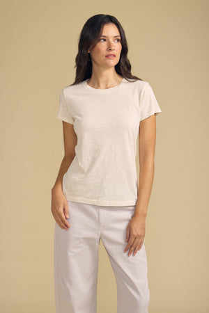 A woman wearing the ODELIA TEE by Velvet by Graham & Spencer—a short-sleeved beige cotton slub top with a classic crew neckline—pairs it with light pants as she stands against a plain beige background, gazing slightly to the side. This wardrobe essential adds understated elegance to any look.