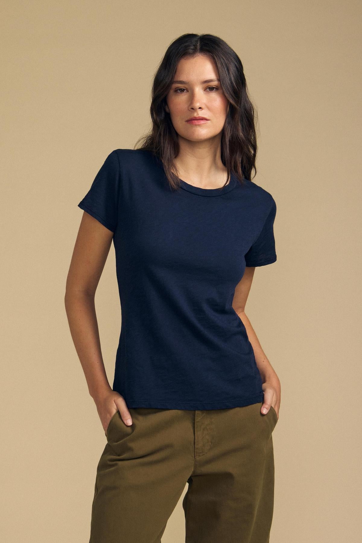 A woman stands against a plain beige background with her hands in her pockets, wearing the Velvet by Graham & Spencer ODELIA TEE in navy blue premium cotton with a classic crew neckline, paired with olive green pants.-41935261040833