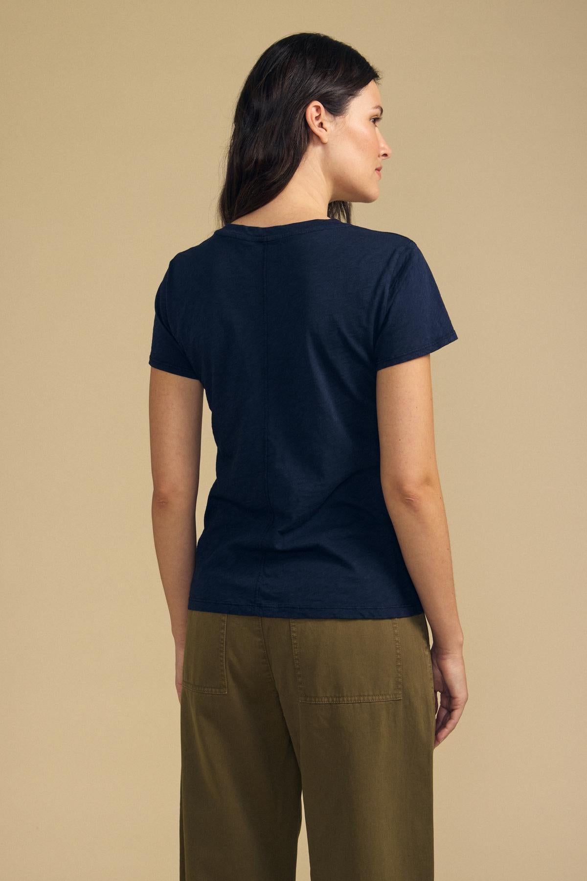 A person with long brown hair stands facing away, wearing the ODELIA TEE by Velvet by Graham & Spencer in classic navy paired with olive green pants against a beige background—a true wardrobe essential.