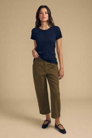 A person stands against a beige background wearing the ODELIA TEE by Velvet by Graham & Spencer—a navy cotton slub tee with a classic crew neckline—paired with olive cropped pants and black flats for a true wardrobe essential look.