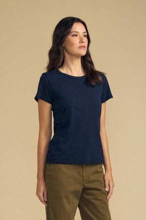 A woman stands against a beige background, wearing the Velvet by Graham & Spencer ODELIA TEE in navy cotton slub with a classic crew neckline, paired with olive green pants and gazing slightly to the side—an effortlessly chic wardrobe essential.