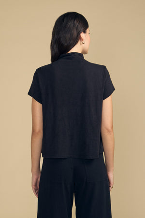A person stands with their back to the camera, wearing the SAVI TEE by Velvet by Graham & Spencer and black pants against a beige background. The top showcases a boxy silhouette for an effortlessly modern look.
