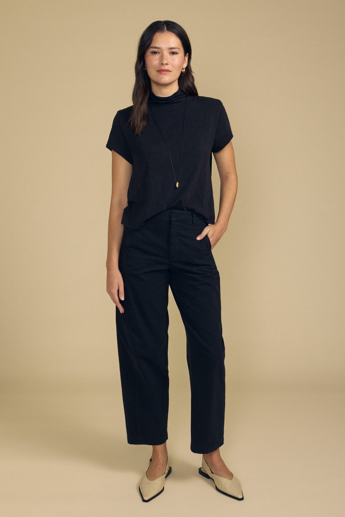 A woman stands against a beige background wearing the SAVI TEE by Velvet by Graham & Spencer—a short-sleeve black cotton slub top with a boxy silhouette—paired with black high-waisted pants, a long necklace, and beige pointed-toe shoes, with one hand in her pocket.
