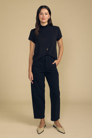 A woman stands against a beige background wearing the SAVI TEE by Velvet by Graham & Spencer—a short-sleeve black cotton slub top with a boxy silhouette—paired with black high-waisted pants, a long necklace, and beige pointed-toe shoes, with one hand in her pocket.