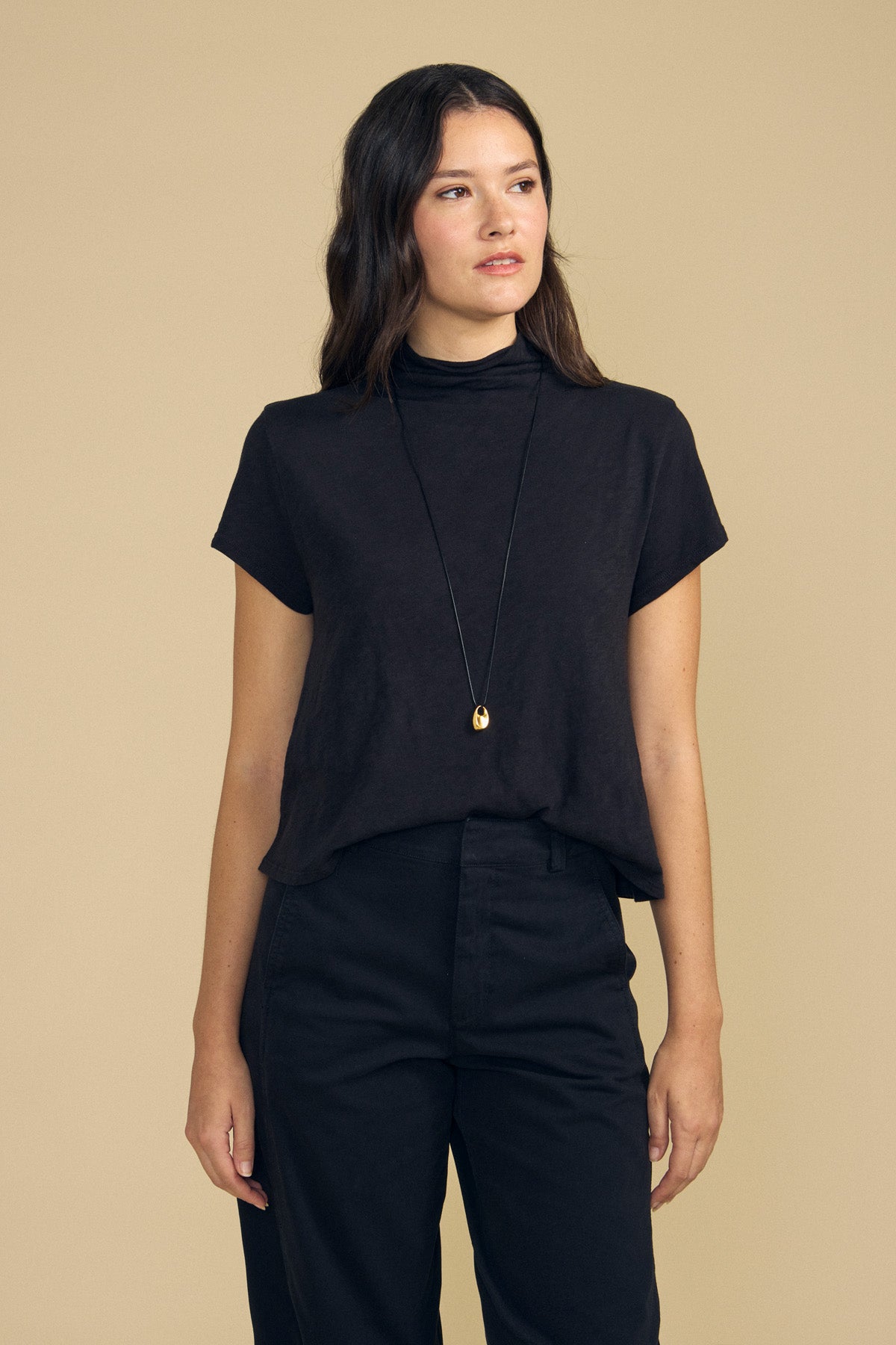 A woman stands against a beige background, wearing the SAVI TEE by Velvet by Graham & Spencer—a black short-sleeve cotton slub top with a boxy silhouette—paired with black pants and a long pendant necklace.