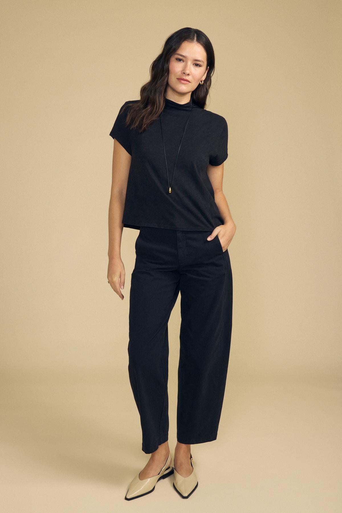 A woman wearing a black short-sleeved mock neck top, Velvet by Graham & Spencer TENLEY PANT high-waisted wide-leg trousers, and pointed-toe beige flats.
