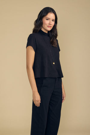 A woman with dark hair and a neutral expression stands against a beige background, wearing the SAVI TEE by Velvet by Graham & Spencer—a black cotton slub tee with a boxy silhouette—styled with dark pants and a long necklace featuring a round pendant.