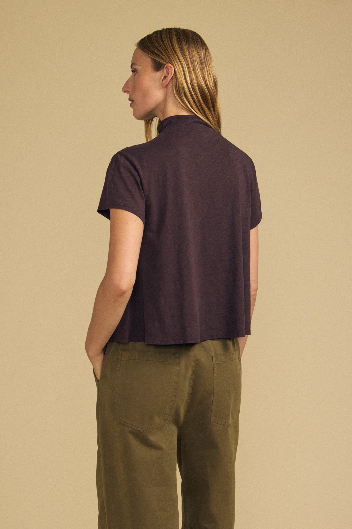 A person with straight, light brown hair stands against a beige background, facing away from the camera, wearing the Velvet by Graham & Spencer SAVI TEE—a short-sleeve dark purple cotton slub tee with a boxy silhouette—paired with olive green pants.