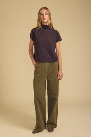 A woman stands against a beige background wearing the SAVI TEE by Velvet by Graham & Spencer—a dark cotton slub top with a boxy silhouette—paired with olive wide-leg pants and brown shoes, one hand in her pocket.