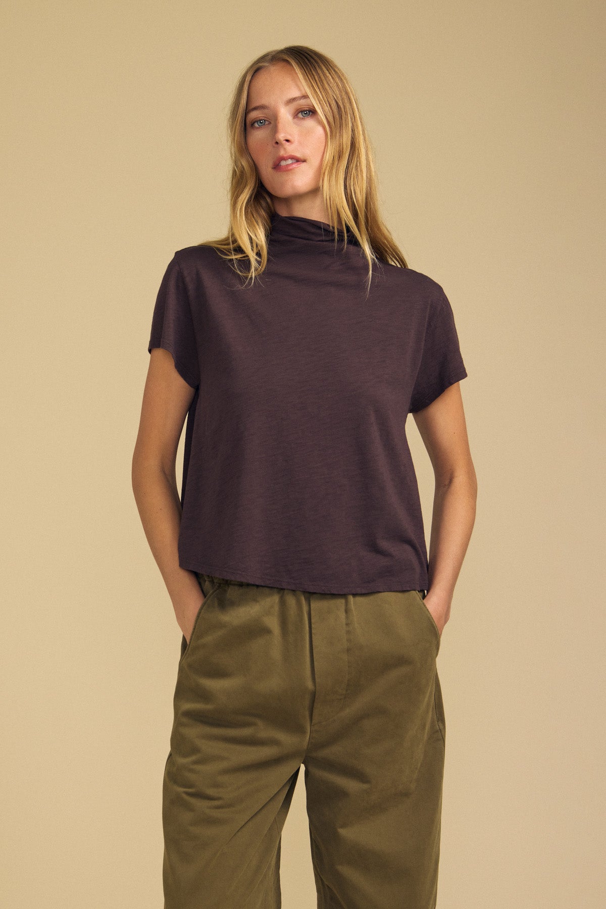 A woman stands with hands in pockets, wearing the SAVI TEE—a relaxed turtleneck short-sleeve shirt by Velvet by Graham & Spencer—paired with high-waisted olive green pants that create a subtle boxy silhouette against a plain beige background.-41929416376513