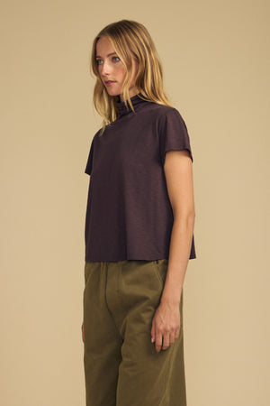A woman is standing sideways against a plain beige background, wearing the SAVI TEE by Velvet by Graham & Spencer—a relaxed, dark purple turtleneck top with a boxy silhouette—paired with olive green pants.