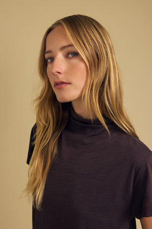 A woman with long blonde hair, wearing the SAVI TEE by Velvet by Graham & Spencer, looks slightly to the side against a plain beige background.