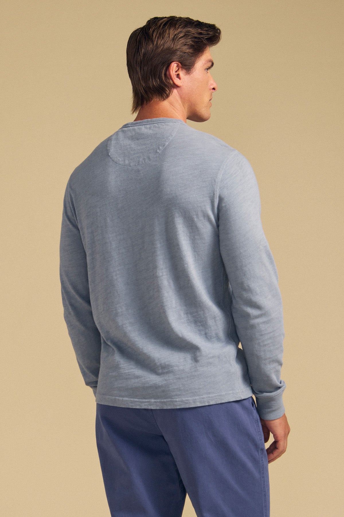 A man faces away, wearing the Velvet by Graham & Spencer ZANE TEE in light blue cotton slub with blue pants, set against a plain beige background.-41548480282817