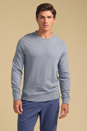 A man faces forward wearing the Velvet by Graham & Spencer ZANE TEE, a light blue cotton slub long sleeve crew neck shirt, and blue pants against a plain beige background.