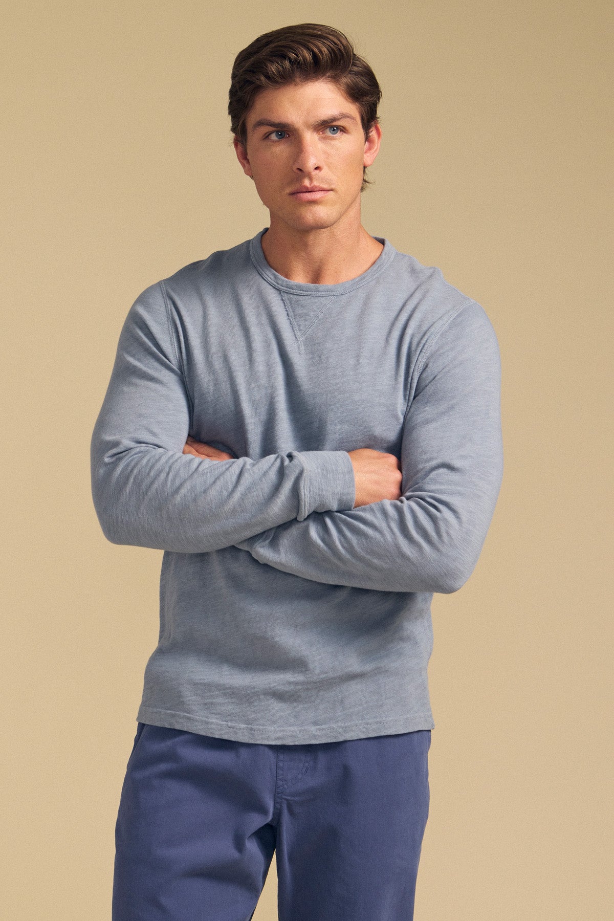 A man with short brown hair stands with arms crossed, wearing the ZANE TEE by Velvet by Graham & Spencer in light blue and blue pants against a plain beige background.-41548480381121