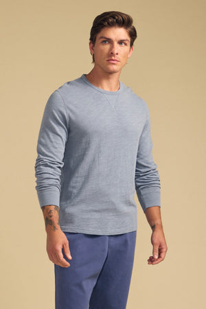 A man wearing the Velvet by Graham & Spencer ZANE TEE in light blue with blue pants stands against a plain beige background.
