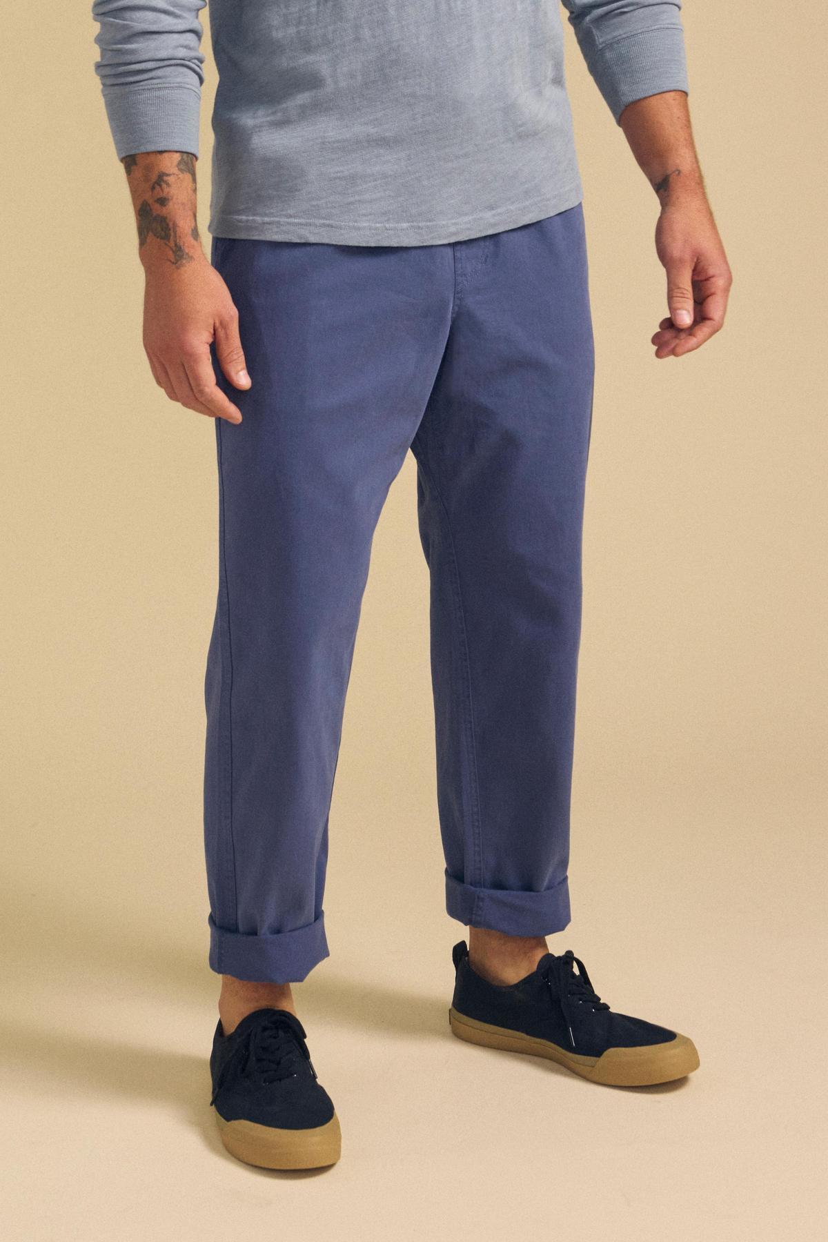 A person wearing the BRANSON PANT by Velvet by Graham & Spencer, featuring blue cuffed legs and an elastic drawstring waist, poses in a light blue long-sleeve shirt and navy sneakers with tan soles against a beige background.-41548481953985