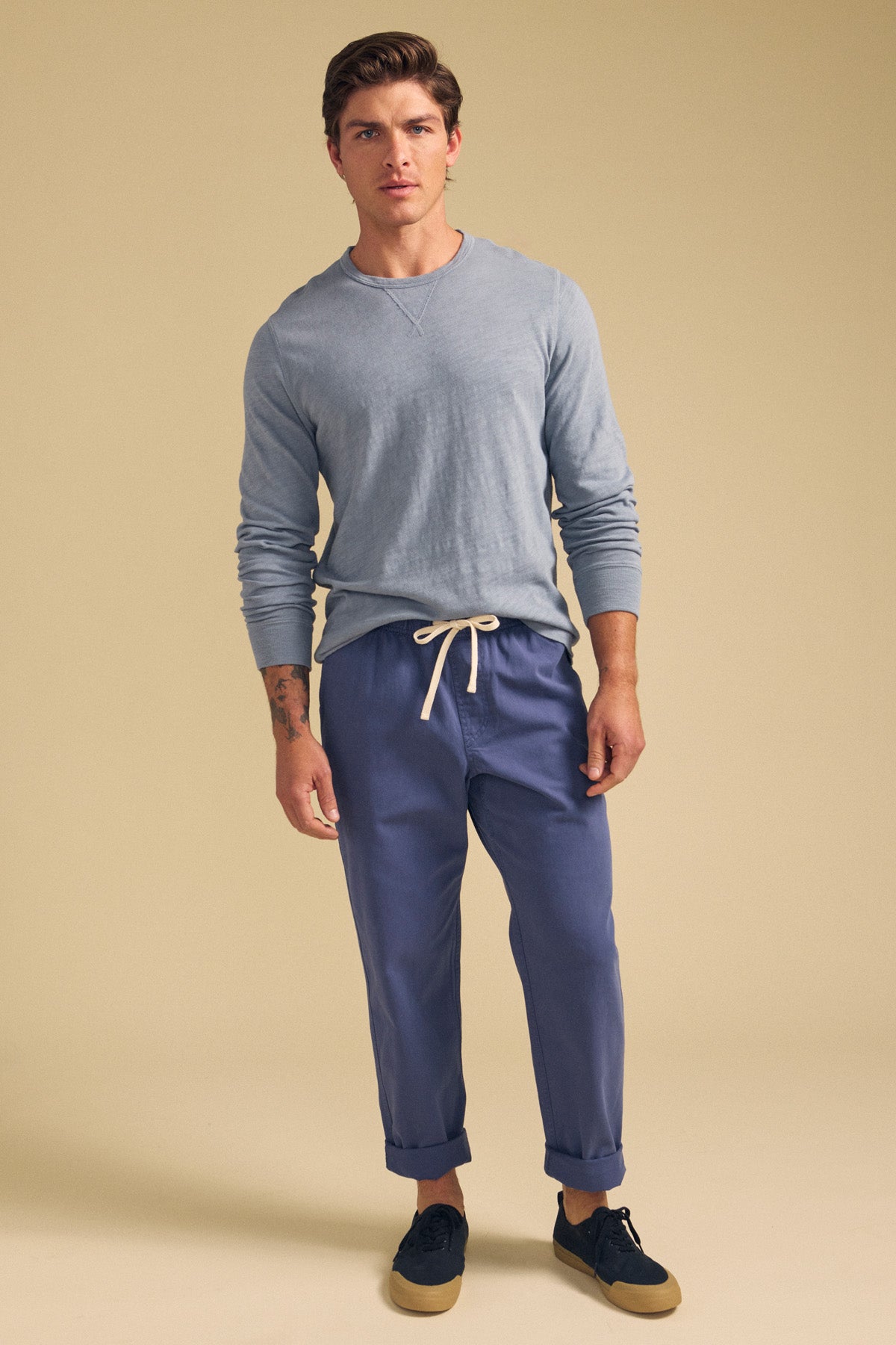 A man faces forward against a plain beige background, wearing the ZANE TEE by Velvet by Graham & Spencer—a light gray long sleeve tee—paired with blue drawstring pants and dark sneakers.-41548480610497