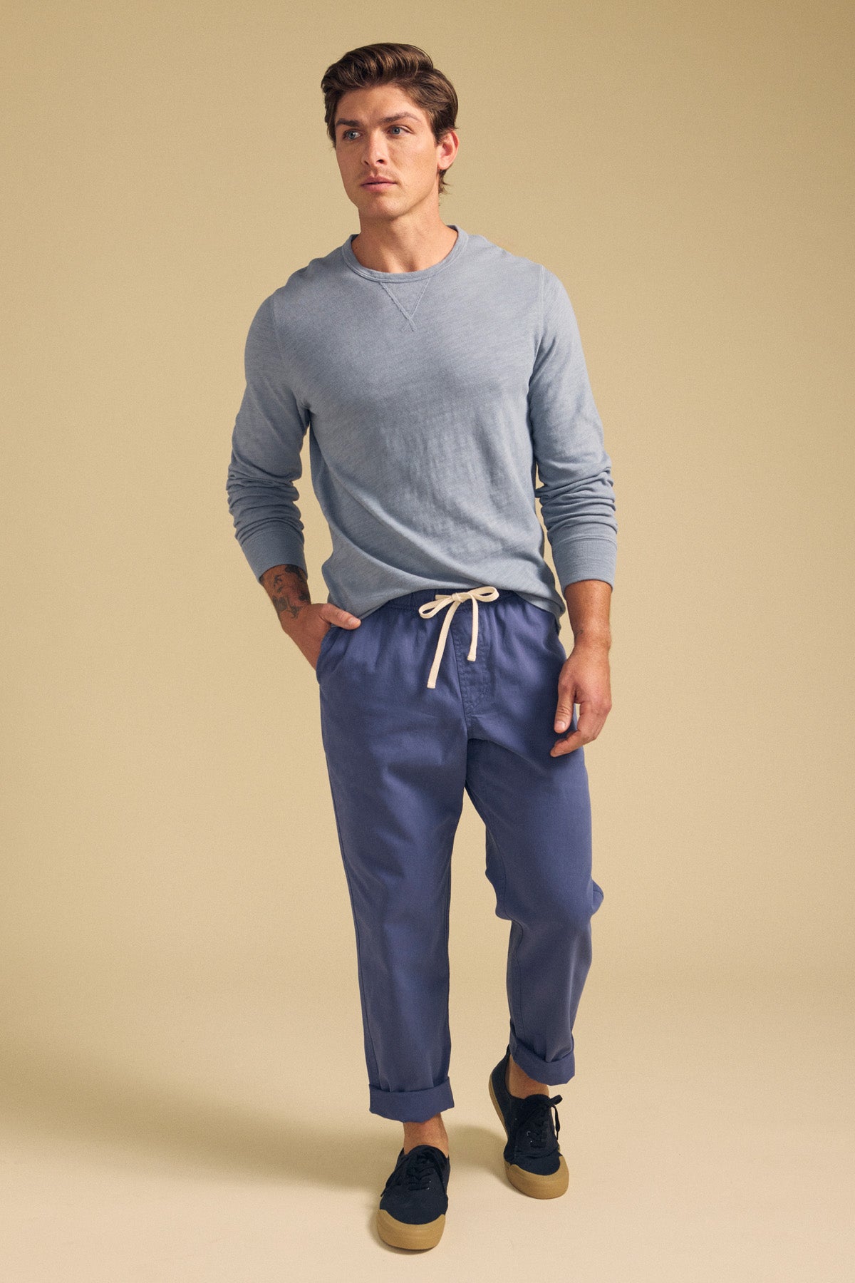 A man stands against a beige background wearing a light blue long-sleeve shirt, BRANSON PANT by Velvet by Graham & Spencer—straight leg blue pants with an elastic drawstring waist—and black sneakers with tan soles.-41548480643265