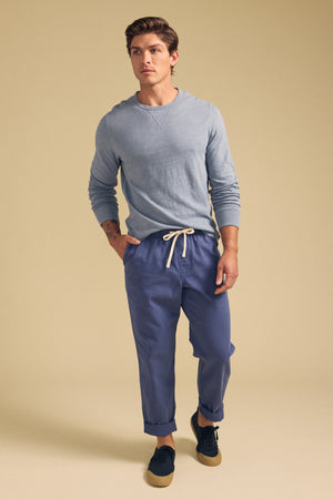 A man stands against a beige background wearing a light blue long-sleeve shirt, BRANSON PANT by Velvet by Graham & Spencer—straight leg blue pants with an elastic drawstring waist—and black sneakers with tan soles.