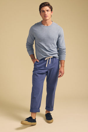 A man stands against a beige background wearing a light blue long-sleeve shirt, Velvet by Graham & Spencer BRANSON PANT with an elastic drawstring waist and rolled cuffs, and black sneakers with tan soles.