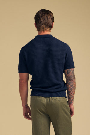 A man faces away, wearing the CARSTON SWEATER POLO by Velvet by Graham & Spencer—a navy cotton knit short sleeve sweater—paired with olive green pants. His right arm shows a sleeve tattoo, set against a plain beige background.